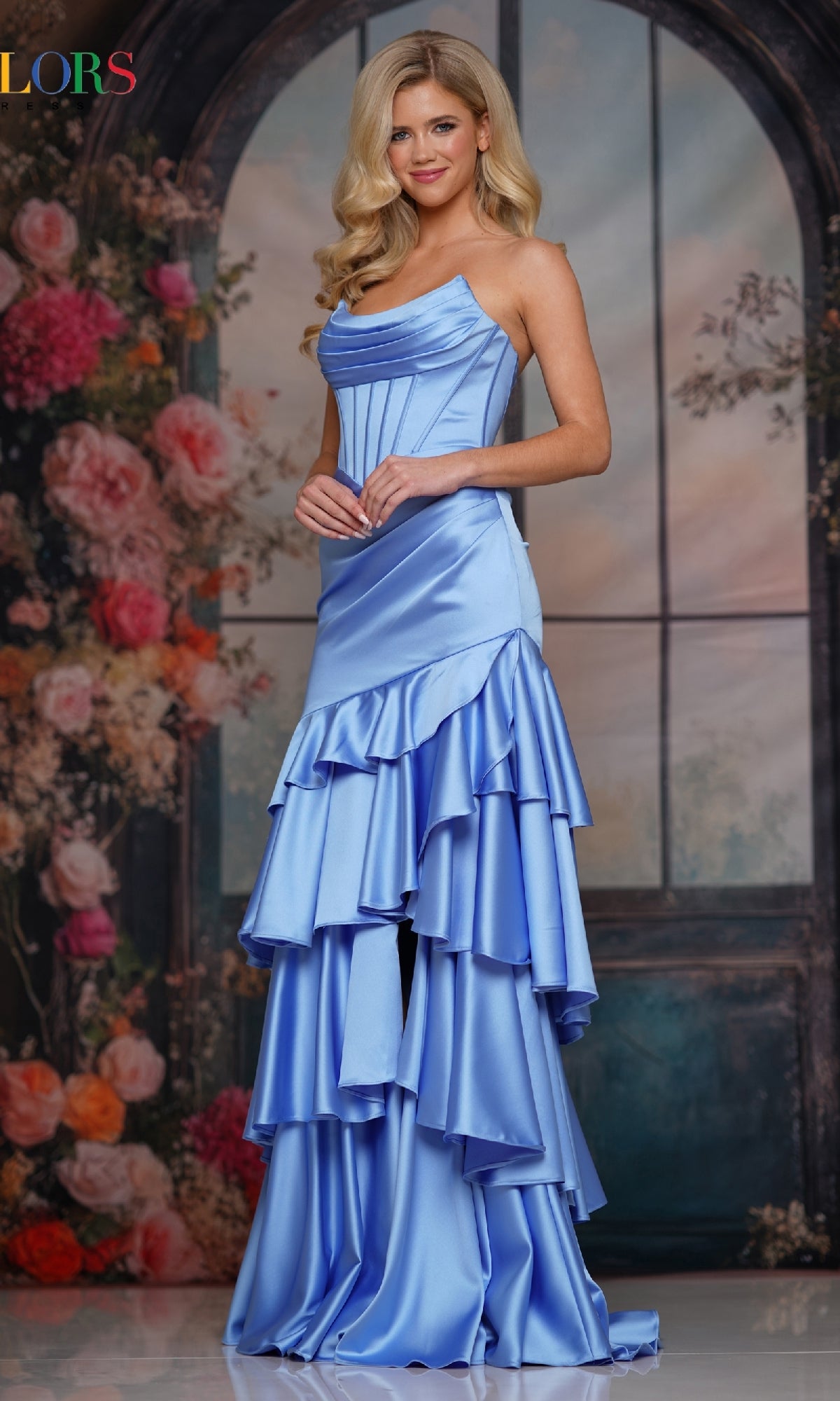 A woman in the Strapless Long Tiered Prom Gown (Colors Dress 3823) in cornflower stands before a floral, arched window backdrop.
